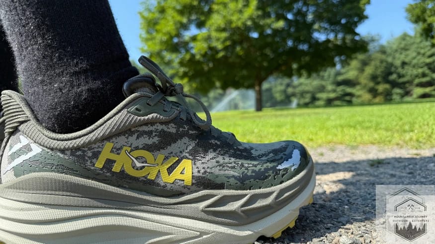The Hoka Stinson 7 on a gravel path with a scenic grass and blue sky background, highlighting its "all-terrain" road-to-trail capabilities.