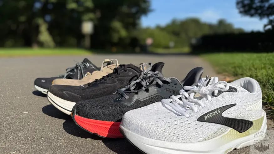 Our 5 best walking shoes lined up on asphalt. From Left to right: Brooks Ghost Max 3 , Altra Experience Flow 2, Hoka Bondi 9, Hoka Transport, Hoka Clifton 10